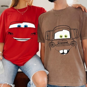 Op de afbeelding: Een felrood t-shirt met een afbeelding van een autogezicht met blauwe ogen en een brede glimlach, en een bruin t-shirt met een afbeelding van een roestig takelwagengezicht met groene ogen, een brede grijns en twee opvallende voortanden op de borst.