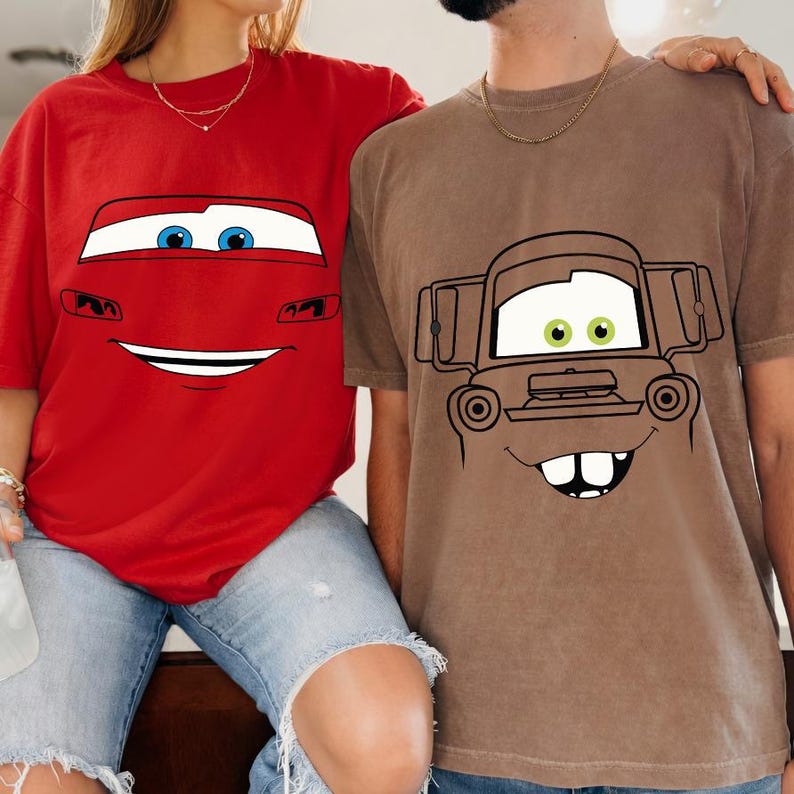 Op de afbeelding: Een felrood t-shirt met een afbeelding van een autogezicht met blauwe ogen en een brede glimlach, en een bruin t-shirt met een afbeelding van een roestig takelwagengezicht met groene ogen, een brede grijns en twee opvallende voortanden op de borst.