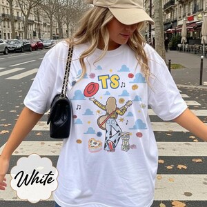 May include: A white t-shirt featuring a graphic print of a person with long hair playing a guitar, wearing a yellow plaid shirt and cow-print pants. The design includes blue clouds, a red hat, musical notes, and blocks with the letters TS.