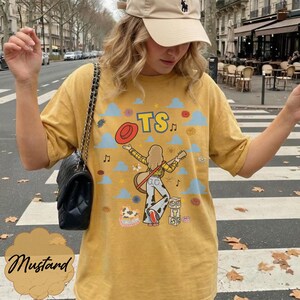 May include: A mustard yellow short-sleeve t-shirt featuring a graphic print of a person with long hair playing a guitar, wearing a yellow plaid shirt and cow-print pants. The design includes blue clouds, a red hat, musical notes, and the text TS.