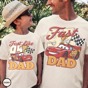 May include: Ivory-colored t-shirts with a red race car graphic, the text "Fast Like 95 Dad", a trophy, and a checkered flag. One shirt is worn by a child, the other by an adult.