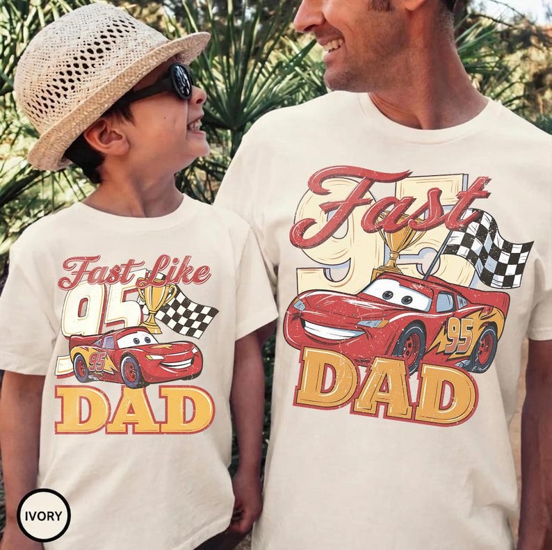 May include: Ivory-colored t-shirts with a red race car graphic, the text "Fast Like 95 Dad", a trophy, and a checkered flag. One shirt is worn by a child, the other by an adult.