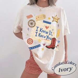 May include: An ivory t-shirt featuring a graphic print of a lined notepad with the text I Knew It, I Knew You, surrounded by illustrations of a red cowboy boot, a sheriff star, a wagon wheel, a target, and various small stars and hearts.