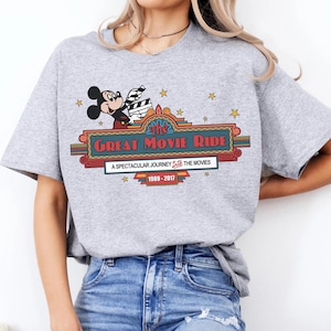 Puede incluir: Camiseta gris claro con un gr&aacute;fico de Mickey Mouse sosteniendo una claqueta de cine sobre las palabras "The Great Movie Ride". El dise&ntilde;o incluye estrellas y el texto "A Spectacular Journey Into The Movies 1989-2017."