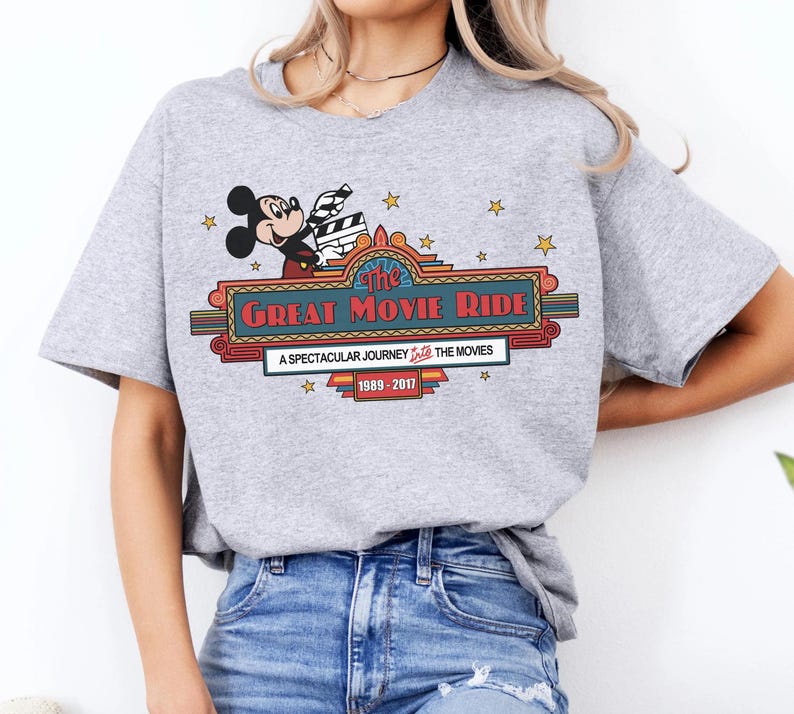 Puede incluir: Camiseta gris claro con un gr&aacute;fico de Mickey Mouse sosteniendo una claqueta de cine sobre las palabras "The Great Movie Ride". El dise&ntilde;o incluye estrellas y el texto "A Spectacular Journey Into The Movies 1989-2017."