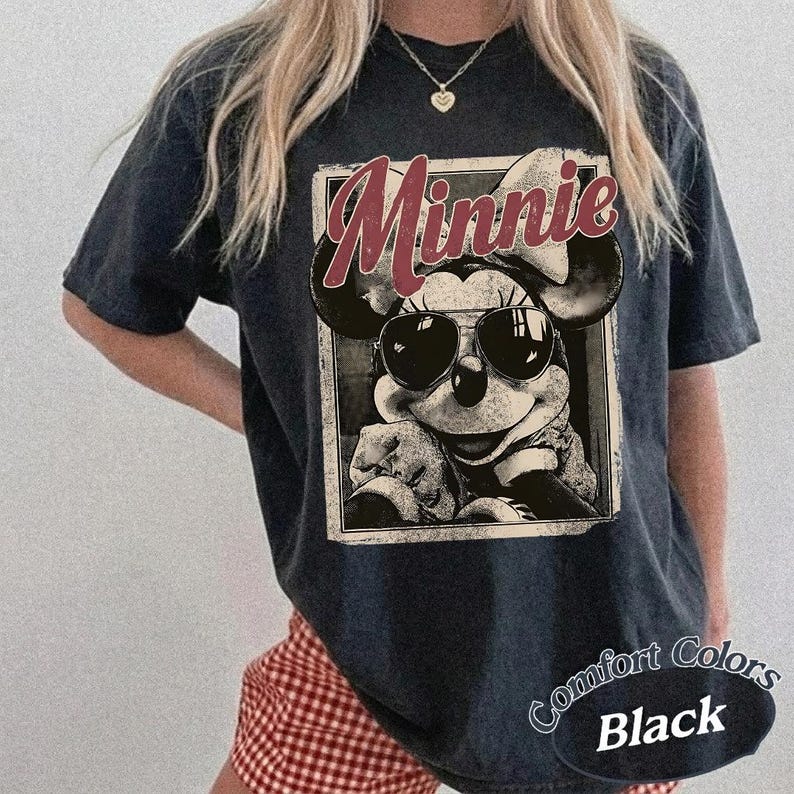 May include: Black t-shirt featuring a graphic of Minnie Mouse wearing sunglasses, with the word "Minnie" in a red, retro font. The shirt has a vintage, distressed look. The words "Comfort Colors Black" are printed on the bottom right.