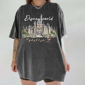 Puede incluir: Camiseta gris oscuro con un dise&ntilde;o de Disneyworld. El dise&ntilde;o incluye un castillo, patos y el texto "Disneyworld" en letra cursiva. La camiseta tiene mangas cortas y un corte holgado.