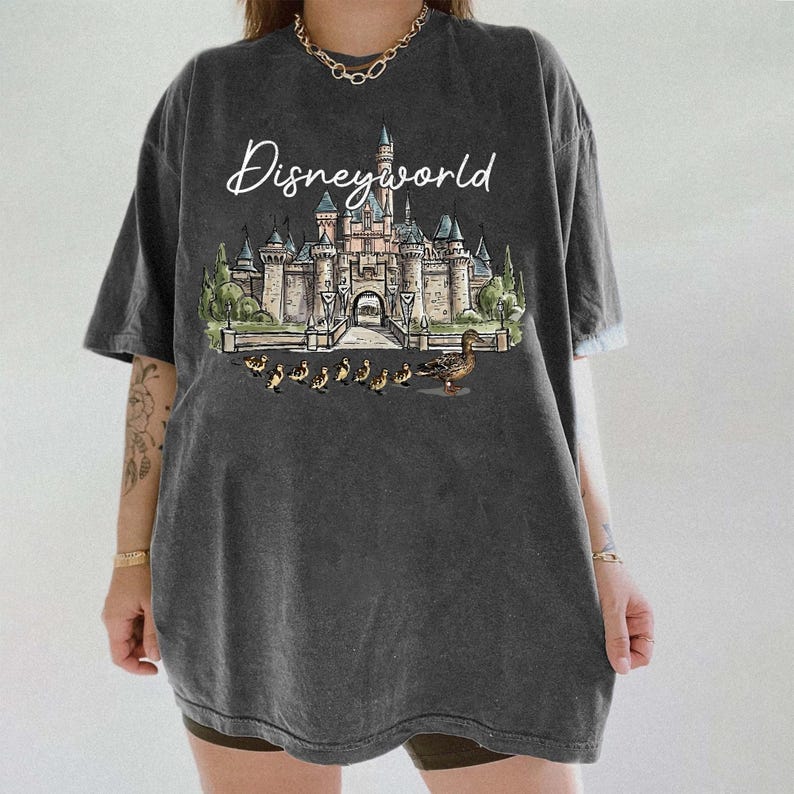 Puede incluir: Camiseta gris oscuro con un dise&ntilde;o de Disneyworld. El dise&ntilde;o incluye un castillo, patos y el texto "Disneyworld" en letra cursiva. La camiseta tiene mangas cortas y un corte holgado.