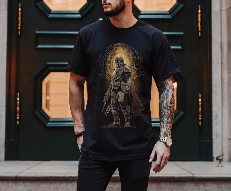 May include: Black t-shirt with a detailed graphic of an armored character set against a golden circular design. The design includes text around the circle. The character is holding a weapon. The shirt has a classic crew neck.