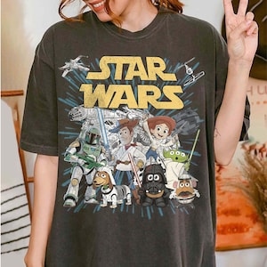 Puede incluir: Camiseta gris oscuro con el texto amarillo "STAR WARS" y un gr&aacute;fico con personajes de Star Wars y Toy Story. El dise&ntilde;o incluye naves espaciales, Darth Vader y otros personajes ic&oacute;nicos.