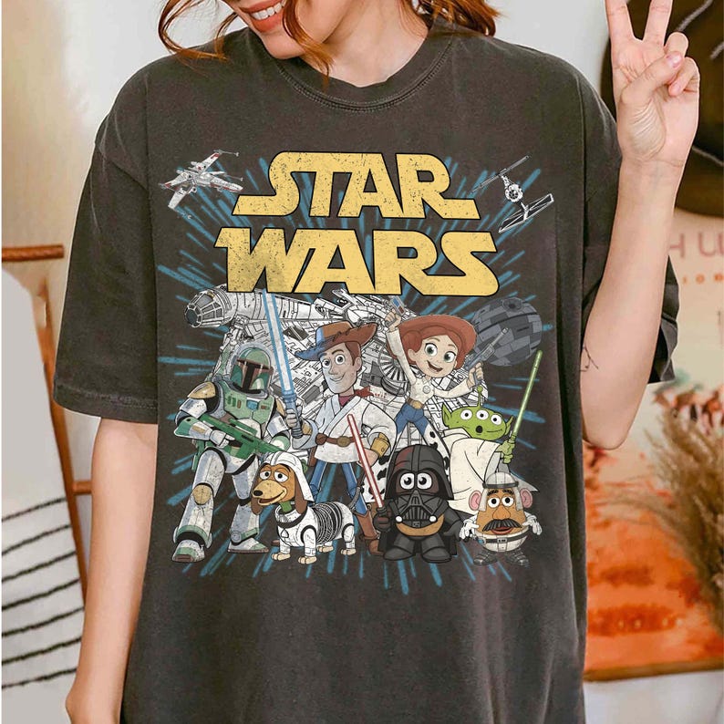 Puede incluir: Camiseta gris oscuro con el texto amarillo "STAR WARS" y un gr&aacute;fico con personajes de Star Wars y Toy Story. El dise&ntilde;o incluye naves espaciales, Darth Vader y otros personajes ic&oacute;nicos.