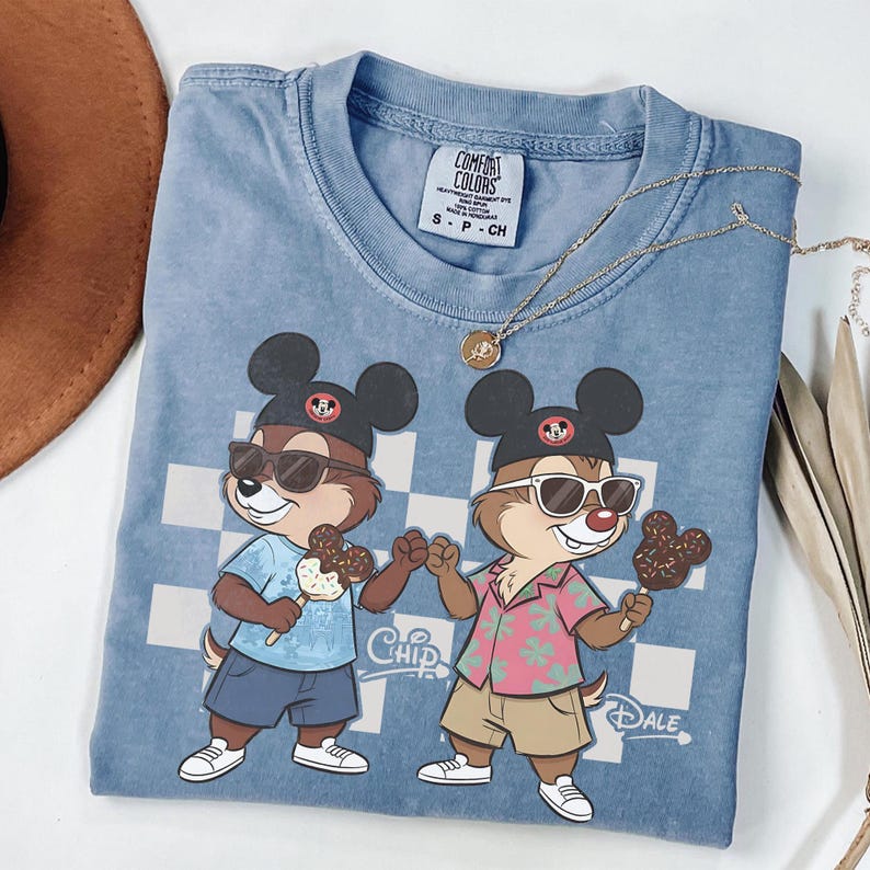 Pu&ograve; includere: Una maglietta blu Comfort Colors con una grafica di Cip e Ciop con occhiali da sole e cappelli di Topolino, che tengono in mano gelati. La maglietta ha uno sfondo a scacchi e una collana d'oro.