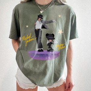 Puede incluir: Camiseta verde oliva con un gr&aacute;fico de Michael Jackson y Mickey Mouse sobre una plataforma morada. El dise&ntilde;o incluye el texto "Michael Jackson" y "Mickey Mouse" en escritura amarilla, con acentos de estrellas.