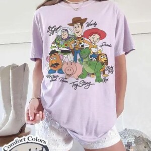 May include: A lilac t-shirt featuring Toy Story characters including Buzz Lightyear and Woody. The words "Toy Story" are printed below the characters. The shirt is a Comfort Colors Orchid.
