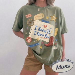 May include: An olive green short-sleeve t-shirt featuring a central graphic of a lined paper scrap with the blue text "I Knew It, I Knew You". The design includes a red cowboy boot, a wagon wheel, a sheriff star, and various small colorful icons.