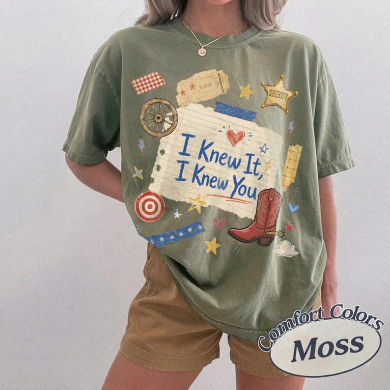 May include: An olive green short-sleeve t-shirt featuring a central graphic of a lined paper scrap with the blue text "I Knew It, I Knew You". The design includes a red cowboy boot, a wagon wheel, a sheriff star, and various small colorful icons.