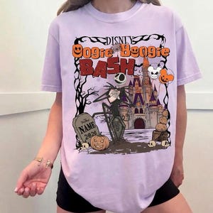 May include: A light purple t-shirt featuring a Halloween-themed graphic. The design includes the text "Disney Oogie Boogie Bash" with Jack Skellington, a castle, pumpkins, and a tombstone. The shirt has short sleeves.