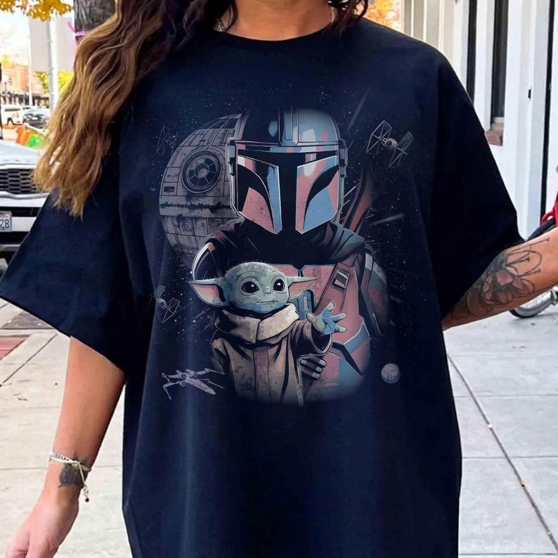 May include: A black t-shirt featuring a graphic print of a Mandalorian character in a helmet standing behind a small green creature with large ears. The background includes a space station, starships, and stars in shades of blue and gray.