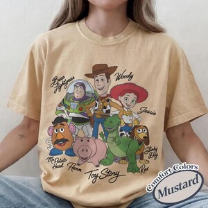 May include: Mustard-coloured t-shirt featuring the Toy Story characters Buzz Lightyear, Woody, Jessie, Mr. Potato Head, Hamm, Rex, and Slinky Dog. The shirt has the words "Toy Story" and "Comfort Colors" printed on it.