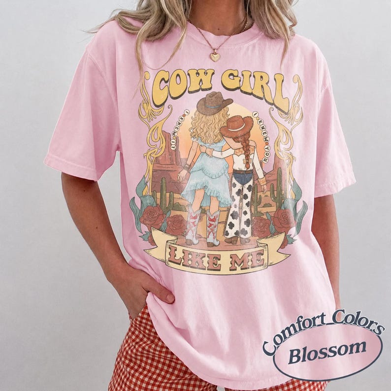 Puede incluir: Una camiseta de manga corta de color rosa claro con un estampado gr&aacute;fico de dos figuras con sombreros de vaquero en un paisaje des&eacute;rtico con cactus y rosas. El texto dice COW GIRL LIKE ME y I KNEW YOU. La camiseta tiene cuello redondo.