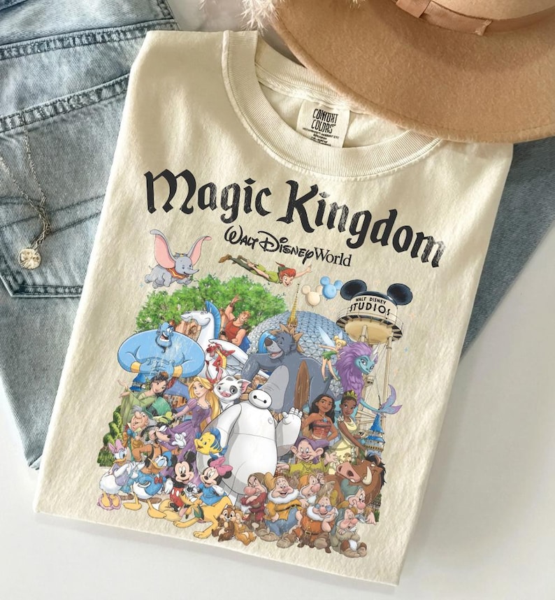 Puede incluir: Camiseta color crema con un gr&aacute;fico de varios personajes de Disney, incluyendo Mickey Mouse, Aladdin y Dumbo. El texto "Magic Kingdom Walt Disney World" est&aacute; impreso en negro en la parte superior. Un sombrero beige y unos vaqueros azules est&aacute;n en el fondo.