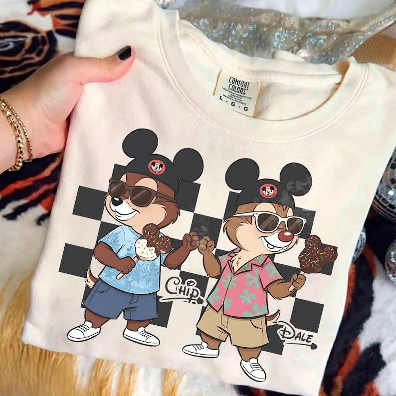 Pu&ograve; includere: Felpa color crema con grafica di Cip e Ciop con occhiali da sole e cappelli di Topolino, che tengono gelati. Il design include uno sfondo a scacchi in bianco e nero e i nomi "Chip" e "Dale".