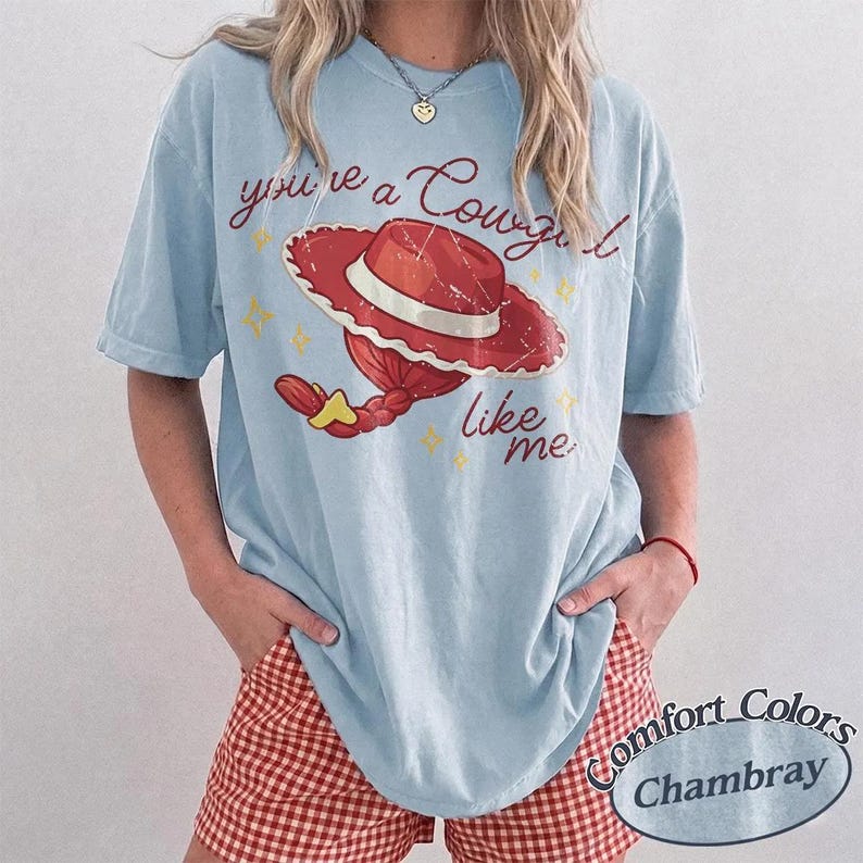 Puede incluir: Una camiseta de manga corta azul claro con un gr&aacute;fico de un sombrero de vaquera rojo y cabello trenzado. El texto dice, you're a Cowgirl like me, acompa&ntilde;ado de peque&ntilde;os iconos de estrellas amarillas. La camiseta tiene un corte holgado.
