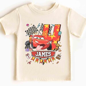 May include: Cream-colored t-shirt featuring a red race car graphic with the number 4, stars, and checkered flags. The name "JAMES" is displayed on a banner below the car. Birthday-themed design.