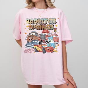 May include: A light pink t-shirt featuring a colorful graphic of characters from the movie "Cars." The design includes the text "Radiator Springs" and "Lightning McQueen" with various car illustrations and stars.