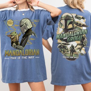K&ouml;nnte beinhalten: Blaues T-Shirt mit einer Grafik von The Mandalorian und Grogu. Das Frontdesign enth&auml;lt den Text "MANDALORIAN" und "THIS IS THE WAY". Das R&uuml;ckendesign zeigt den Text "THE MANDALORIAN AND GROGU" und andere Star Wars-Bilder.