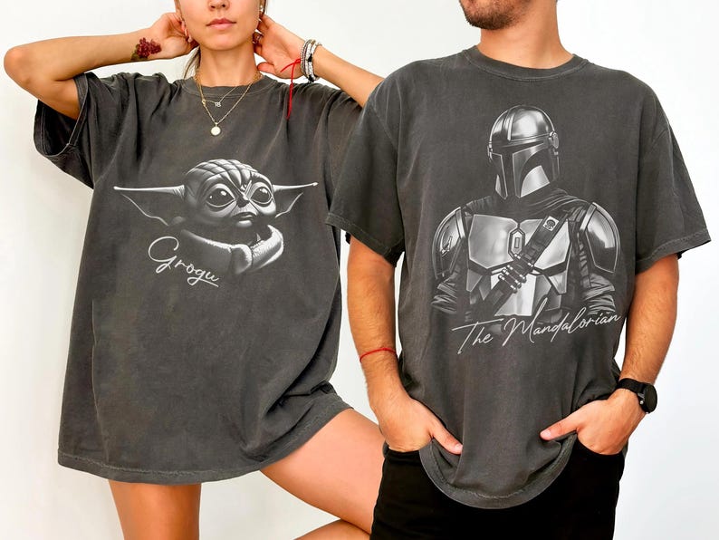 May include: Two charcoal gray t-shirts. One features a Grogu graphic with the name "Grogu" in cursive. The other displays a detailed illustration of The Mandalorian with the text "The Mandalorian" below the image.