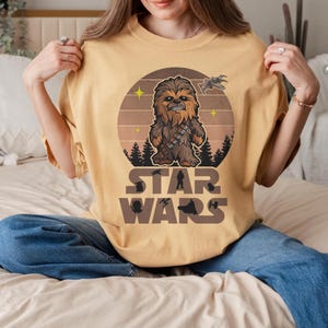 Op de afbeelding: Mosterdgeel T-shirt met een Chewbacca-afbeelding in een cirkelvormig ontwerp. Het ontwerp bevat een ruimteschip, sterren en de woorden "STAR WARS" in een bruin lettertype. Het shirt is gemaakt van een zacht materiaal.