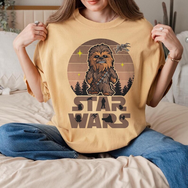 Op de afbeelding: Mosterdgeel T-shirt met een Chewbacca-afbeelding in een cirkelvormig ontwerp. Het ontwerp bevat een ruimteschip, sterren en de woorden "STAR WARS" in een bruin lettertype. Het shirt is gemaakt van een zacht materiaal.