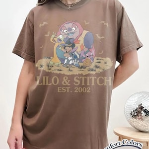 May include: Espresso-colored t-shirt featuring a graphic of Lilo & Stitch characters in Halloween costumes. The text "Lilo & Stitch EST. 2002" is printed above the characters. The shirt is from Comfort Colors.