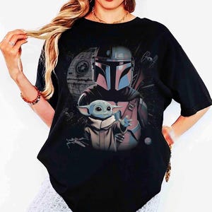May include: A black short-sleeved t-shirt featuring a graphic print of a Mandalorian character in a helmet standing behind a small green creature with large ears. The background includes a space station, starships, and stars in a dark cosmic scene.