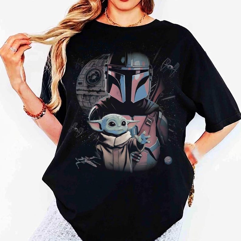 May include: A black short-sleeved t-shirt featuring a graphic print of a Mandalorian character in a helmet standing behind a small green creature with large ears. The background includes a space station, starships, and stars in a dark cosmic scene.
