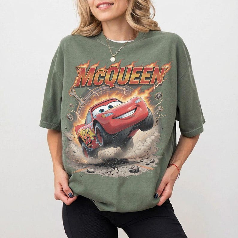 Op de afbeelding: Olijfgroen T-shirt met een afbeelding van Bliksem McQueen uit de film Cars. Het ontwerp bevat het woord "McQueen" in een vlammende lettertype boven de rode raceauto. Het shirt heeft korte mouwen en een relaxte pasvorm.