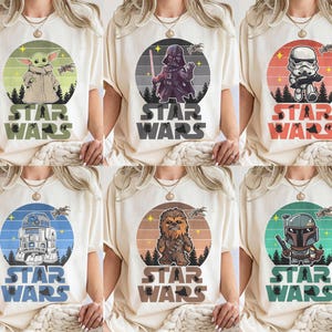 Op de afbeelding: Cr&egrave;mekleurige T-shirts met Star Wars-personages: Baby Yoda, Darth Vader, een Stormtrooper, R2-D2, Chewbacca en een Mandalorian. Elk ontwerp bevat een karakterillustratie in een cirkelvormige afbeelding en de woorden "STAR WARS".
