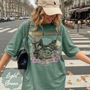 Puede incluir: Camiseta verde claro con un gr&aacute;fico de The Mandalorian, Grogu y una nave espacial, rodeados de flores. La palabra "Mandalorian" est&aacute; impresa en la parte superior. La camiseta es de color verde claro.