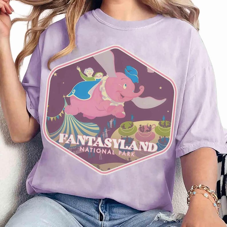 Op de afbeelding: Een lichtpaars t-shirt met een zeshoekige grafische print van een vliegende roze olifant boven een circustent en een theekopjesattractie. Het ontwerp bevat de tekst FANTASYLAND NATIONAL PARK in witte letters op een donkerpaarse achtergrond.