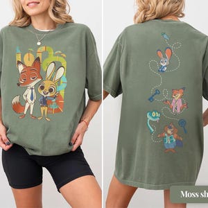 May include: Moss green t-shirt featuring the characters Nick Wilde and Judy Hopps from Zootopia. The front of the shirt displays the characters, while the back showcases other characters and objects from the movie. The shirt is made of a soft material.