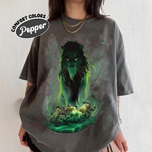 Puede incluir: Camiseta gris oscuro Comfort Colors con una ilustraci&oacute;n verde y negra de un le&oacute;n y tres hienas. La camiseta tiene el texto "Comfort Colors Pepper" en un dise&ntilde;o circular.