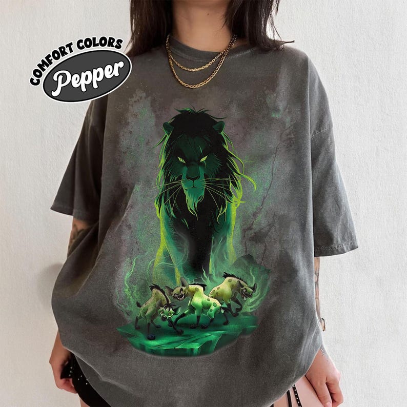 Puede incluir: Camiseta gris oscuro Comfort Colors con una ilustraci&oacute;n verde y negra de un le&oacute;n y tres hienas. La camiseta tiene el texto "Comfort Colors Pepper" en un dise&ntilde;o circular.