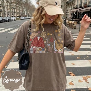 May include: A brown short-sleeve t-shirt featuring a graphic print of a cowboy hat, a red paisley cardigan, a sheriff badge labeled Sheriff Taylor, a microphone, a guitar, and blocks spelling Toy Story, alongside a black shoulder bag.