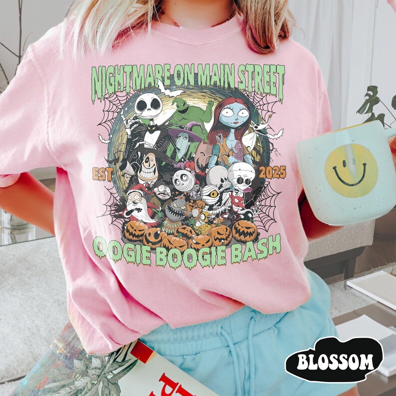 Puede incluir: Sudadera rosa con un dise&ntilde;o gr&aacute;fico de personajes de "Pesadilla antes de Navidad" y el texto "Nightmare on Main Street OOGIE BOOGIE BASH 2025". Se sostiene una taza azul claro con una carita sonriente amarilla.