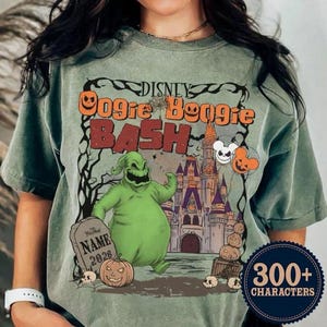 May include: Sage green t-shirt featuring a graphic for the Disney Oogie Boogie Bash. The design includes the text "Disney Oogie Boogie Bash" in orange and red, with a cartoon character, pumpkins, and a castle. The shirt has a crew neck and short sleeves.