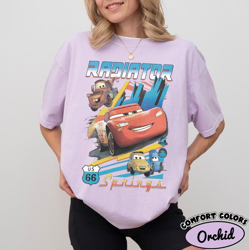 May include: A light purple t-shirt featuring a graphic print of characters from the movie Cars. The design includes the text Radiator Springs and a US 66 road sign. The print shows a red race car, a brown tow truck, and two smaller vehicles.