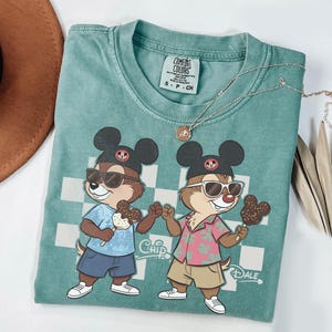 Pu&ograve; includere: Una maglietta Comfort Colors verde acqua con una grafica di Cip e Ciop con occhiali da sole e gelato. La maglietta ha uno sfondo a scacchi e le parole "Chip" e "Dale" sono stampate. Una collana d'argento &egrave; drappeggiata sulla maglietta.