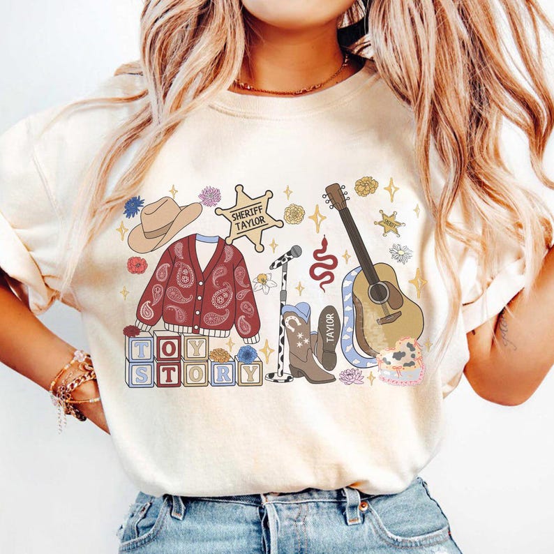 May include: A cream-colored t-shirt featuring a graphic print of a cowboy hat, a paisley cardigan, a sheriff star badge labeled Sheriff Taylor, a microphone, a guitar, cowboy boots, and blocks spelling Toy Story with various floral accents.