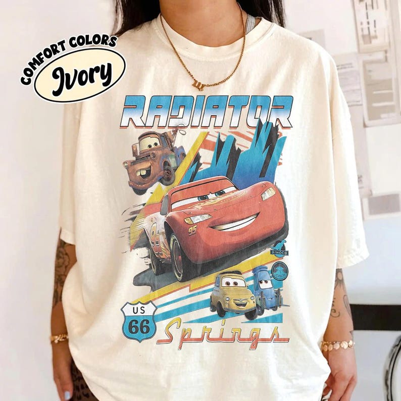 May include: An ivory t-shirt featuring a graphic print of characters from the movie Cars, including Lightning McQueen and Mater. The text reads Radiator Springs and US 66. The shirt has a crew neckline and short sleeves, with a Comfort Colors Ivory label.